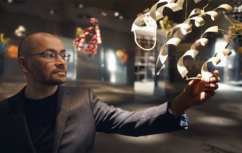 AI-onderzoeker Demis Hassabis in de documentaire ‘The Thinking Game’ Man grijpt in een museumachtige omgeving naar een uitvergrote eiwitstructuur