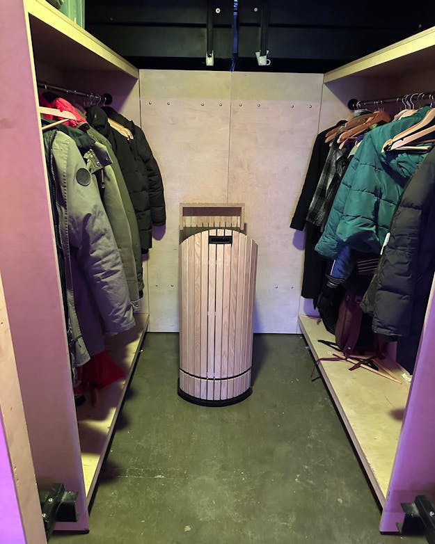 Robotober Rober staat in de garderobe tussen de jassen. Een garderoberuimte met links en rechts jassen aan hangers In het midden tegen de achtermuur staat iets wat op een prullenbak lijkt.