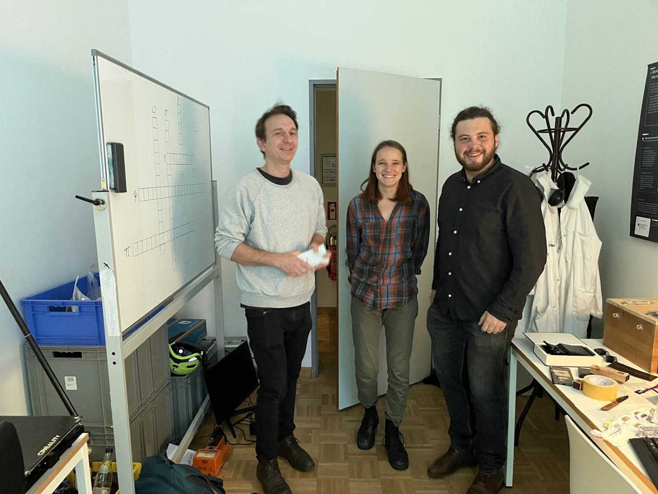 Drie promovendi poseren in de door hun bedachte quantum-escaperoom. In de kamer staat een bureau, een whiteboard en een kapstok waar witte labjassen aan hangen.