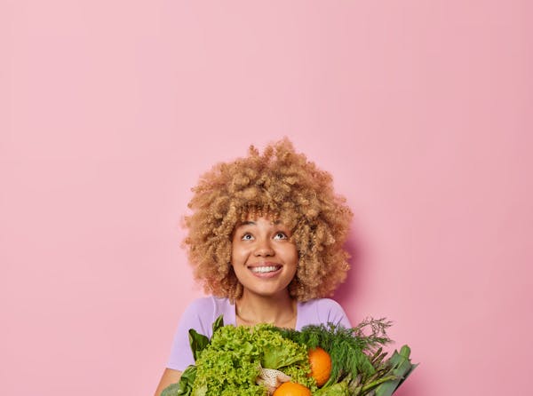 jonge vrouw met springerig haar die heel veel verschillende groenten vasthoudt en blij kijkt