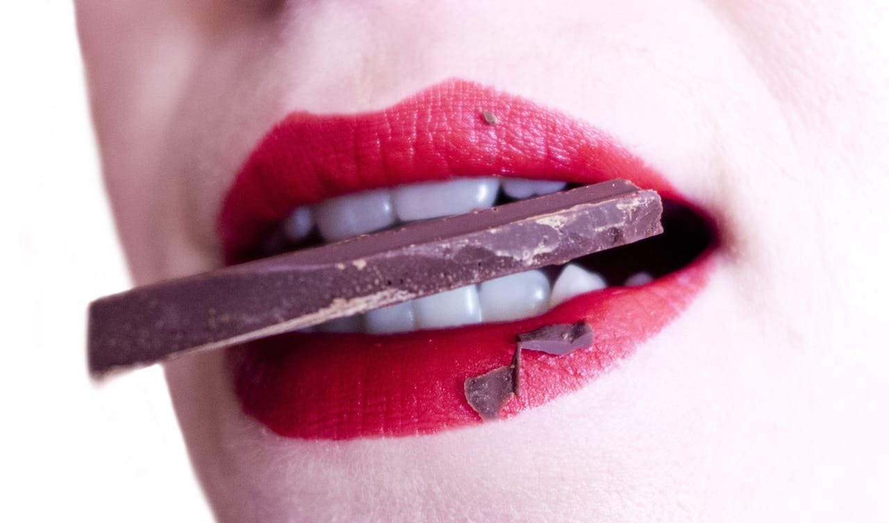 Een close-up van een mond met rode lippenstift die op een stuk chocolade bijt.