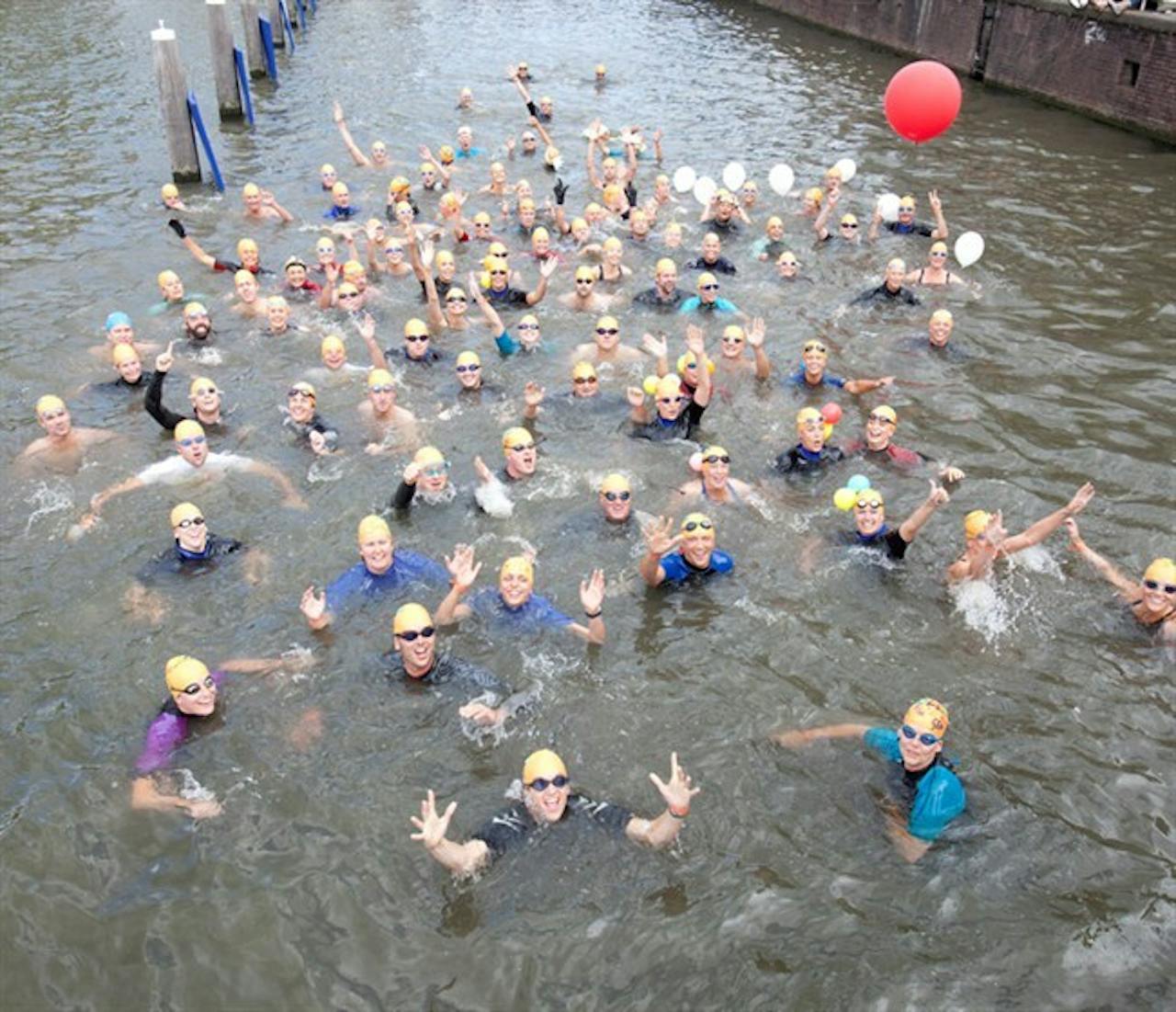 Een groep mensen die zwemmen in een gracht. Op de achtergrond is een rode ballon te zien.