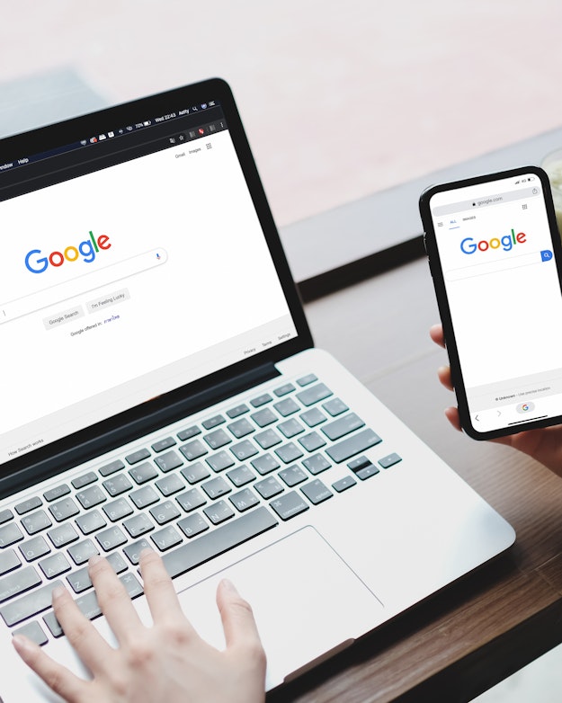 Een vrouw heeft een laptop opengeklapt voor zich staan met op het scherm zoekmachine Google. In haar rechterhand houdt ze haar mobiele telefoon, waarop ook Google is geopend.