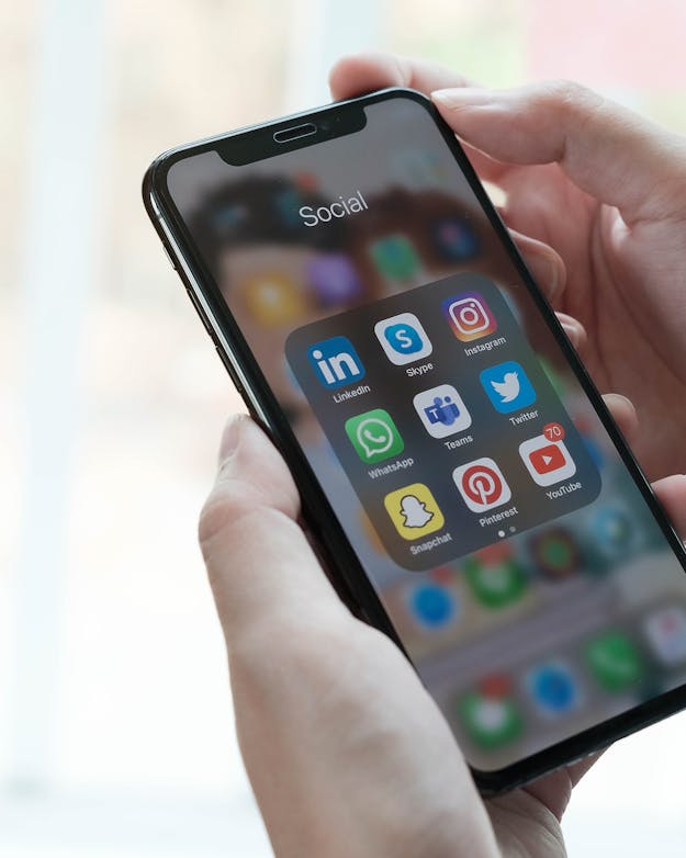Een vrouwenhand houdt een iPhone vast. Op het scherm is een mapje 'Social' te zien, meer daarin de iconen van verschillende socialmedia-apps. Een vrouwenhand houdt een iPhone vast. Op het scherm is een mapje 'Social' te zien, meer daarin de iconen van verschillende socialmedia-apps.