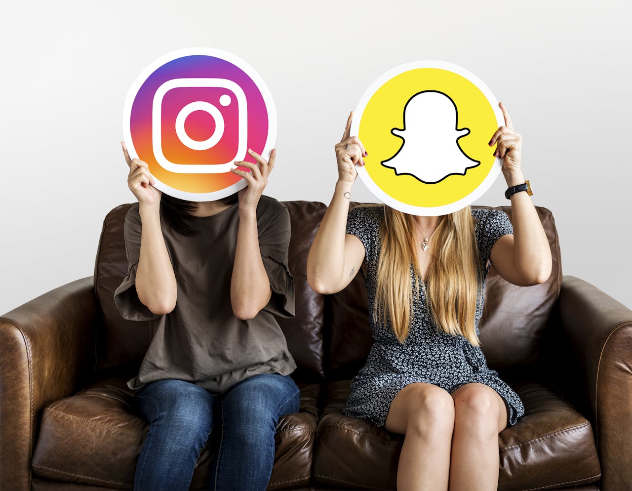 Twee vrouwen zitten op een bank. De ene houdt het logo van Instagram voor haar gezicht, de andere het logo van Snapchat.