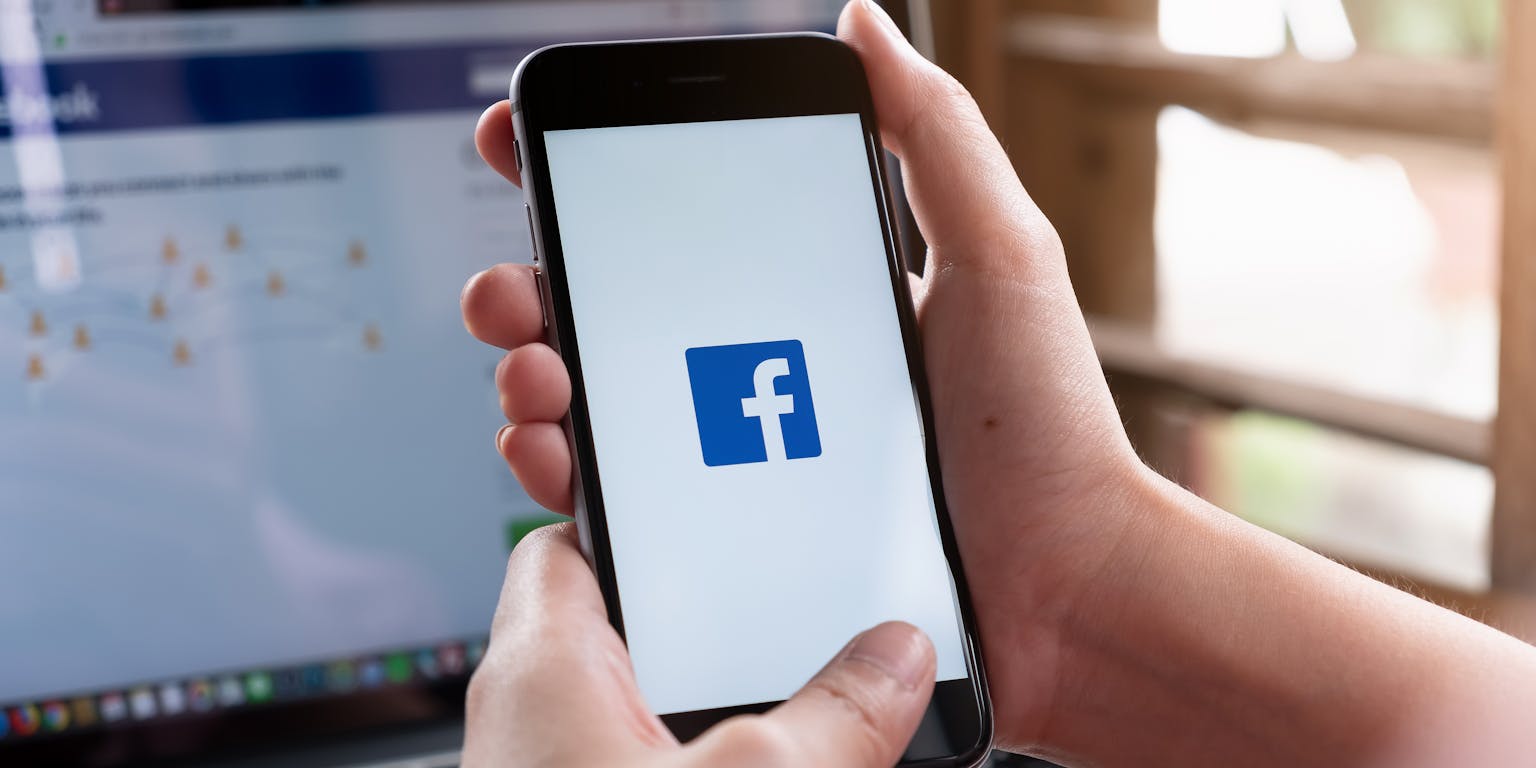 Twee vrouwenhanden houden een smartphone vast met het logo van Facebook op het scherm. Op de achtergrond is een laptop te zien met het inlogscherm van Facebook in beeld.