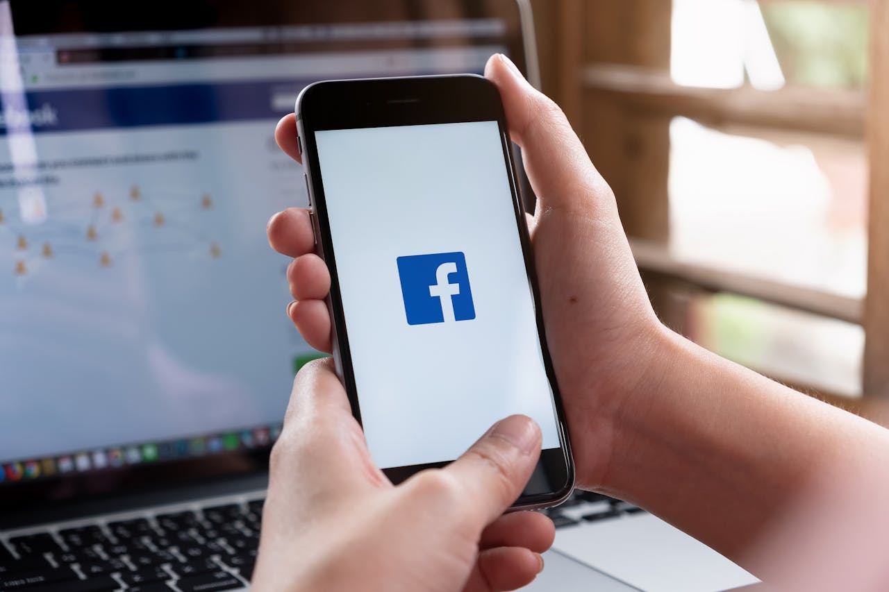 Twee vrouwenhanden houden een smartphone vast met het logo van Facebook op het scherm. Op de achtergrond is een laptop te zien met het inlogscherm van Facebook in beeld.