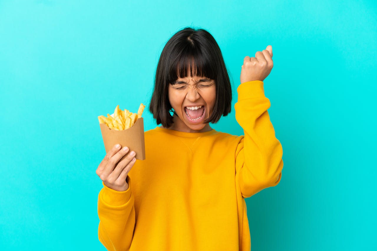 tiener met gele trui en bruin haar en lichtblauwe achtergrond maakt overwinningsgebaar met bakje patat in haar hand