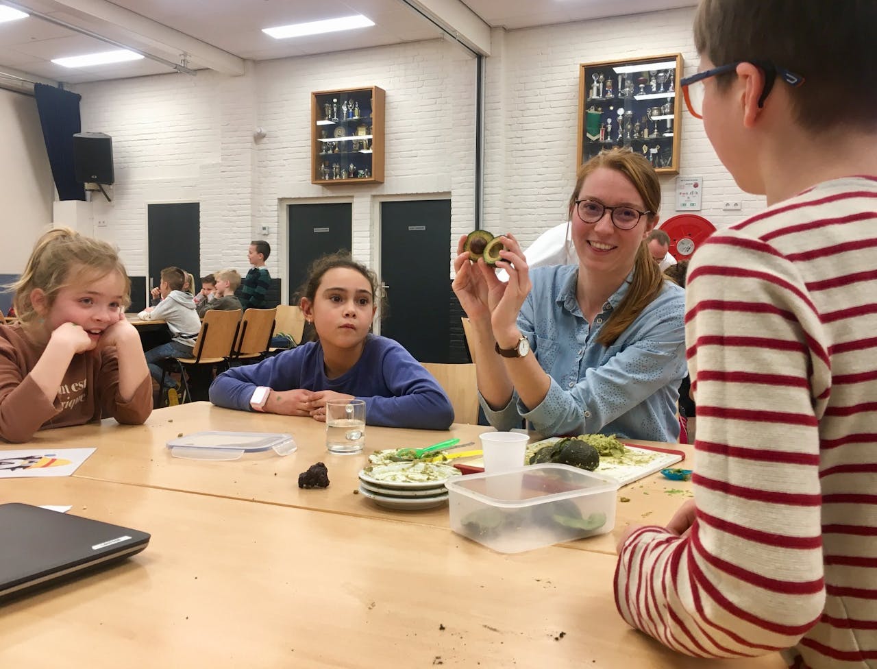 Een groep kinderen en een volwassene in een klaslokaal. De volwassene houdt een avocado in de lucht.